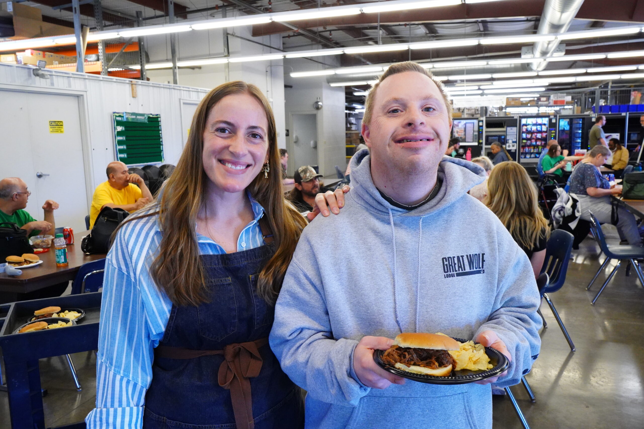 Staff member stands beside a participant holding a plate with a sandwich and chips during a group meal.