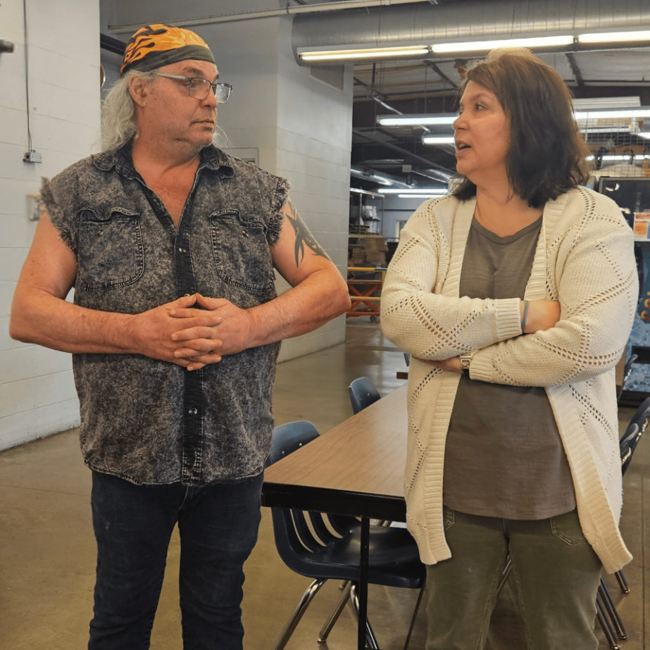 Two people talk with each other while standing near tables in a dining and activity space.